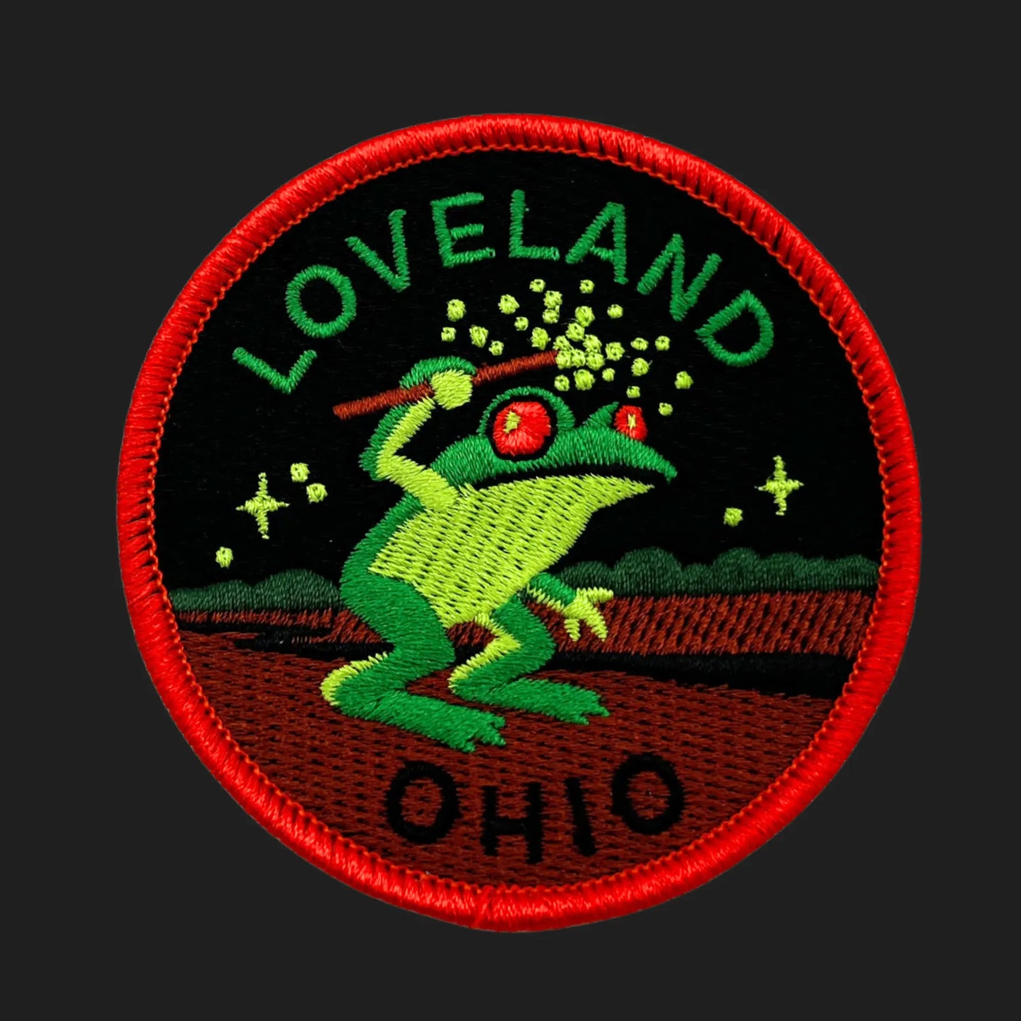 Embroidered patch depicting the Loveland Frogman of Ohio — a green humanoid frog with glowing red eyes holding a wand emitting sparks, set against a black night sky with stars and trees, bordered in bright red with the words “LOVELAND” and “OHIO.”