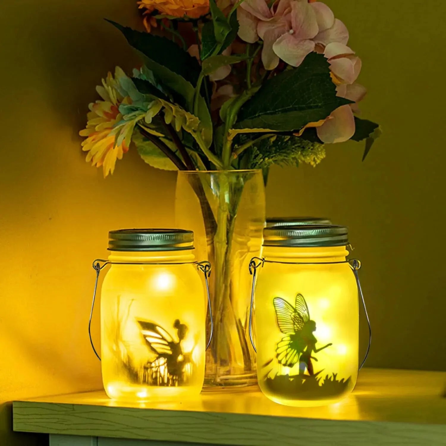 Two glowing fairy solar lanterns with yellow light on a table beside a glass vase of colorful flowers