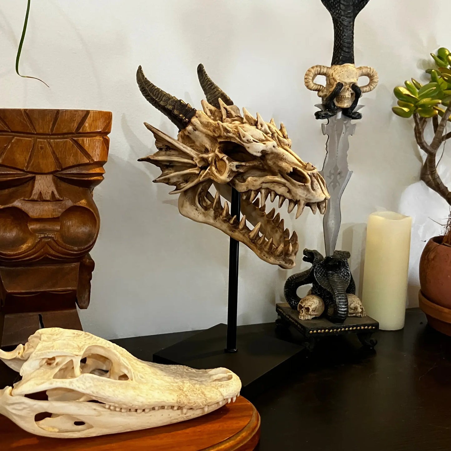 Dragon skull replica on stand next to tiki carving, dagger, and crocodile skull, styled as a gothic fantasy display.