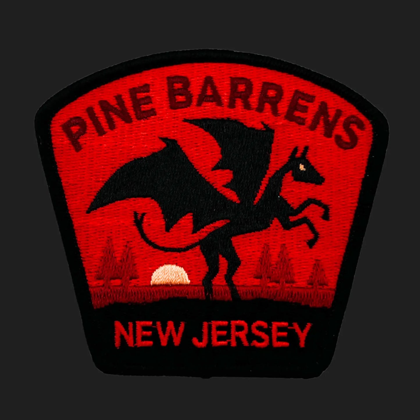 Embroidered patch showing the Jersey Devil — a winged, horned creature rearing on its hind legs — set against a red sunset background with pine trees. Text reads “PINE BARRENS” above and “NEW JERSEY” below in bold red lettering.