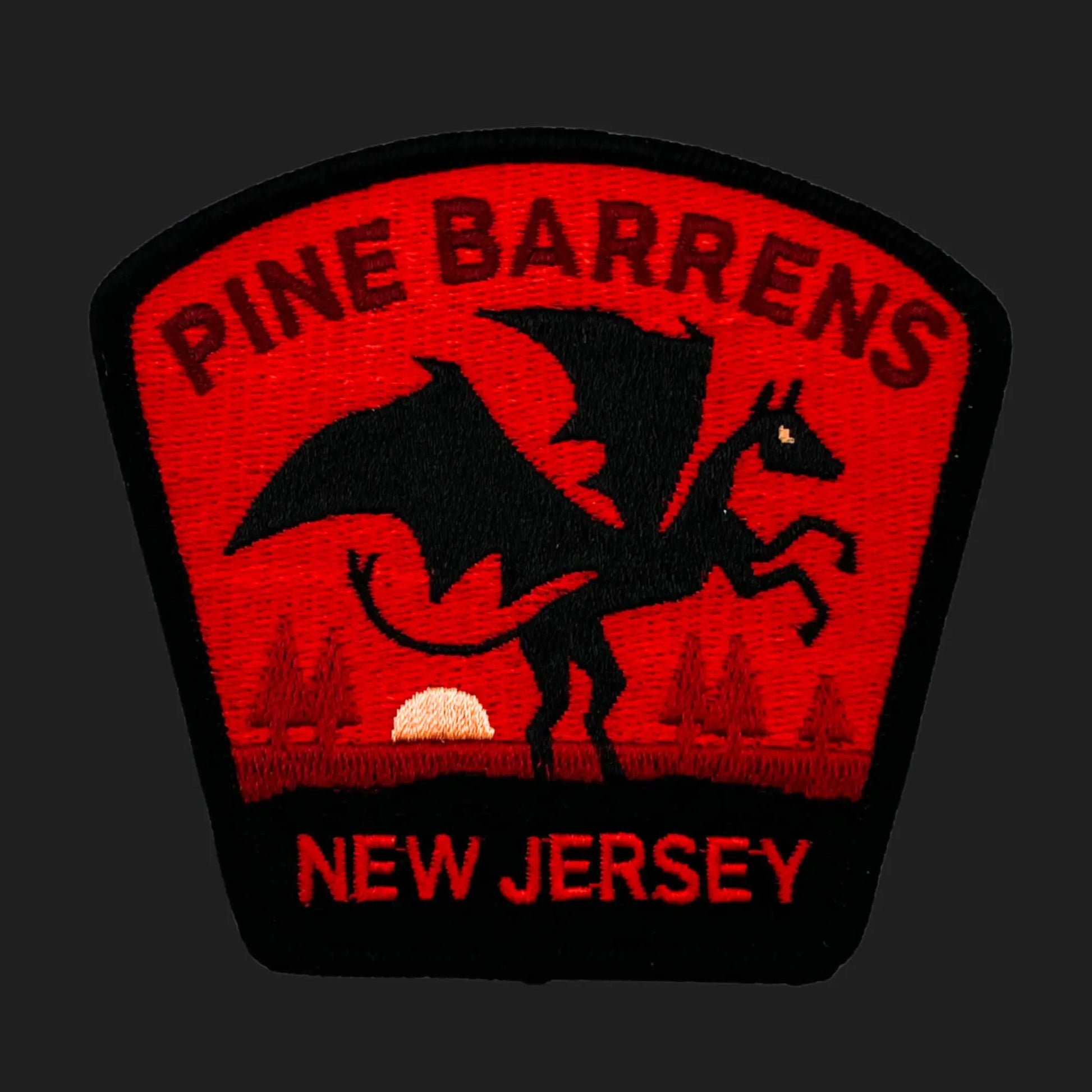 Embroidered patch showing the Jersey Devil — a winged, horned creature rearing on its hind legs — set against a red sunset background with pine trees. Text reads “PINE BARRENS” above and “NEW JERSEY” below in bold red lettering.