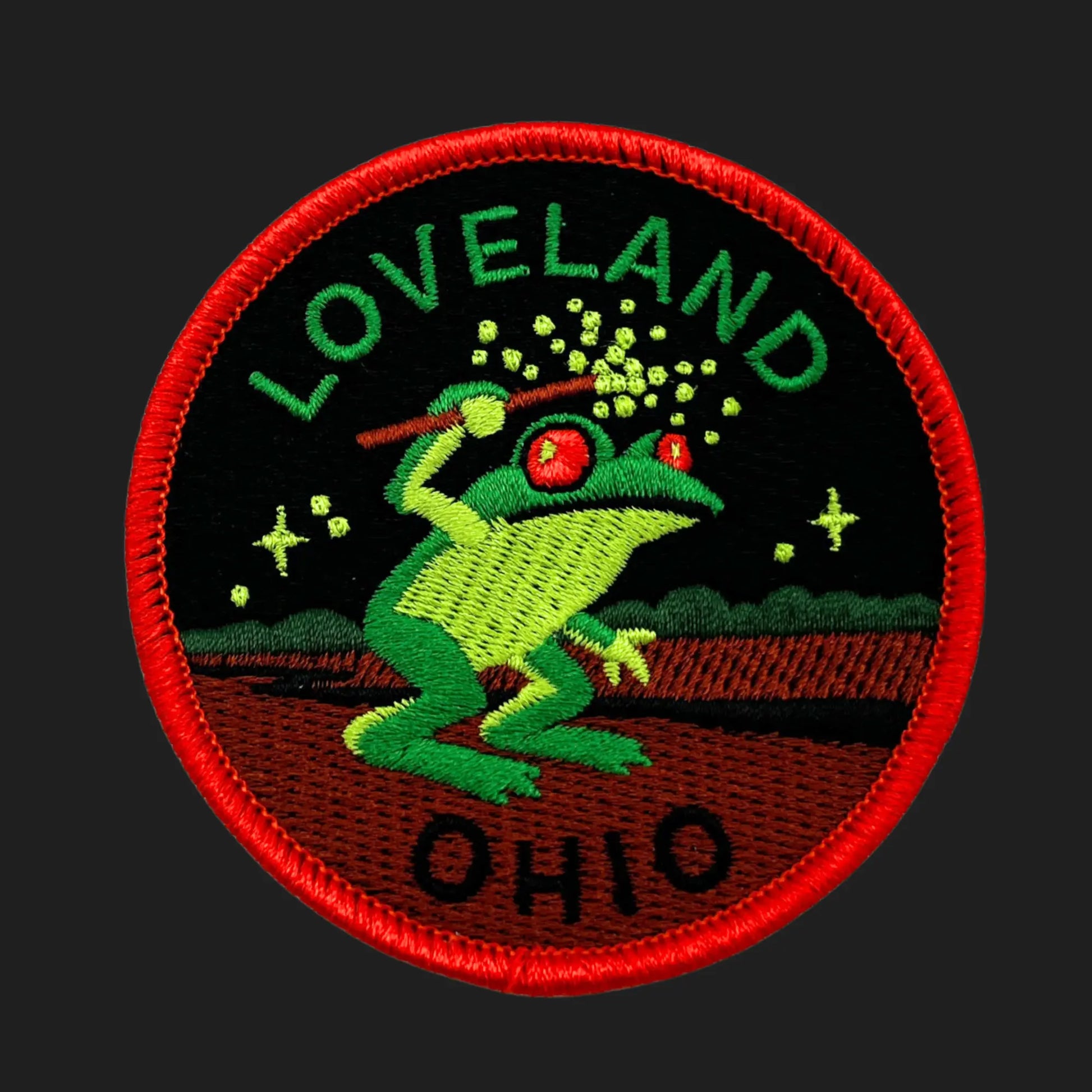 Embroidered patch depicting the Loveland Frogman of Ohio — a green humanoid frog with glowing red eyes holding a wand emitting sparks, set against a black night sky with stars and trees, bordered in bright red with the words “LOVELAND” and “OHIO.”