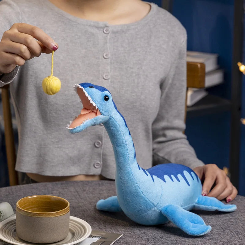Person playing with blue Nessie plush toy using a yellow yarn ball, seated at a table with a ceramic cup