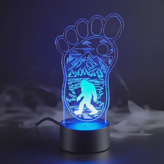 Bigfoot footprint LED lamp cycling through multiple colors with etched mountain, forest, and Sasquatch design.