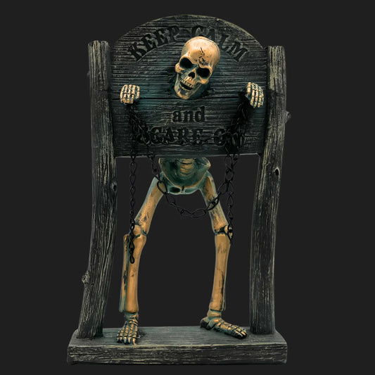 Skeleton statue in wooden stocks with chains, engraved with 'Keep Calm and Scare On'