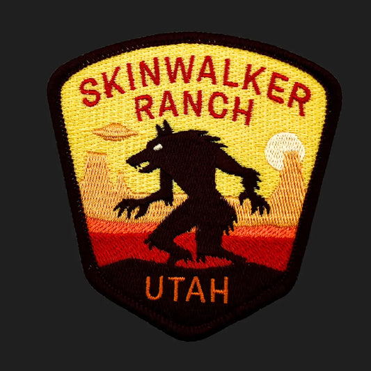 Embroidered patch featuring a silhouette of the Skinwalker creature against a desert sunset background with mesas, a glowing sun, and a UFO in the sky. The design reads “SKINWALKER RANCH” and “UTAH” in red and orange thread.